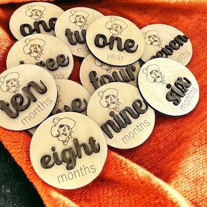 May include: Wooden monthly milestone markers, each with a cowboy illustration and a number or time period like "one year" or "six months". The markers are displayed on orange fabric. The measurements are in months.