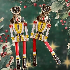 Puede incluir: Un par de pendientes de cascanueces de tono dorado con detalles de esmalte rojo, blanco, amarillo y negro. Los cascanueces llevan sombreros negros con detalles dorados.