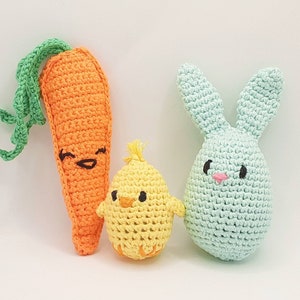 May include: Three crocheted stuffed toys: a green bunny, an orange carrot, and a yellow chick. The bunny has a pink nose and black eyes. The carrot has a smiling face. The chick has black eyes and a beak.