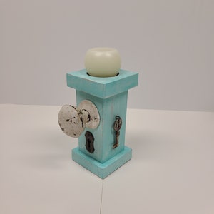 May include: A teal-painted wooden candle holder with a vintage door knob, key, and keyhole accents. A white candle sits atop the square structure. The candle holder is a unique decorative item.