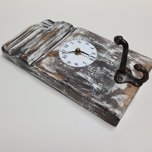 May include: A rustic wooden wall clock with a white face and black hands. The clock is mounted on a piece of reclaimed wood with a distressed white paint finish. A black metal hook is attached to the side of the wood.