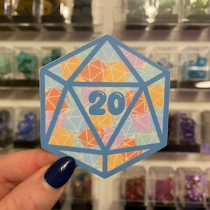 Pode incluir: Um autocolante azul em forma de dado de vinte lados, com o número 20 no centro. O dado é preenchido com um padrão geométrico colorido de formas laranja, rosa e azul. O autocolante é segurado em frente a uma exibição de pequenos objetos coloridos.