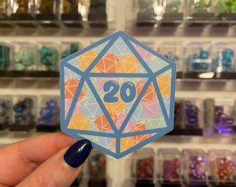 May include: A blue sticker in the shape of a twenty-sided die, with the number 20 in the centre. The die is filled with a colourful geometric pattern of orange, pink, and blue shapes. The sticker is held in front of a display of small, colourful objects.