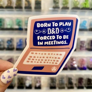 Puede incluir: Una pegatina con forma de portátil rosa con el texto "BORN TO PLAY D&D FORCED TO BE IN MEETINGS" en una pantalla azul. El fondo está borroso, mostrando estantes con objetos coloridos.