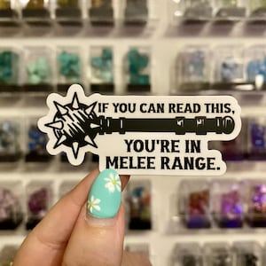 May include: A white sticker featuring a black mace graphic and text that says, "IF YOU CAN READ THIS, YOU'RE IN MELEE RANGE." The sticker is held by a hand with a turquoise nail with white flower designs.