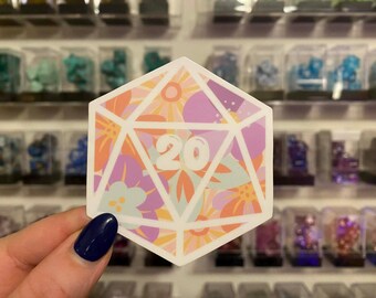 May include: A colorful D20 sticker with a floral design in shades of orange, purple, and light blue. The number 20 is prominently displayed in white. The sticker is held in front of a blurred background of small display cases.