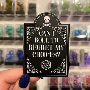 Peut inclure: Autocollant noir avec texte blanc : « CAN I ROLL TO REGRET MY CHOICES ? » avec un crâne et des os croisés, et un symbole de dé. L'autocollant a des ornements décoratifs et est tenu devant une vitrine de minéraux colorés.
