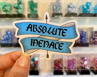 May include: A sticker with the words "ABSOLUTE MENACE" in black script on a light blue banner. The banner is outlined in white with a black arrow through it. The background features various dice sets in display cases.