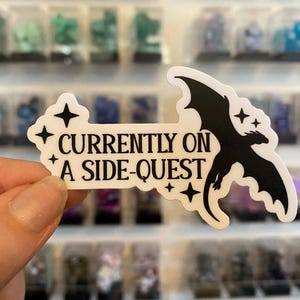 May include: A white sticker featuring the text "CURRENTLY ON A SIDE-QUEST" and a black dragon illustration. The sticker has a star design and is held in front of a display of small, colourful objects.