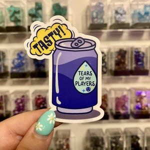 Puede incluir: Una pegatina con un diseño de lata de refresco de dibujos animados en tonos morados y azules. La lata tiene el texto "TEARS OF MY PLAYERS" en una etiqueta y un bocadillo que dice "TASTY!"