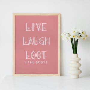 May include: A framed print with the words "LIVE LAUGH LOOT (THE BODY)" in white, set against a dusty rose background. The print is in a light wood frame. A white vase with white flowers is next to the print.