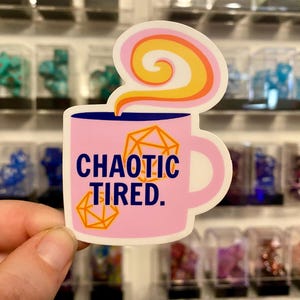 Könnte beinhalten: Eine rosa Kaffeetasse mit blauem Rand und einem gelben und weißen Dampfdesign. Die Tasse hat einen weißen Henkel und sagt "CHAOTIC TIRED." mit einer rosa und orangefarbenen Würfelgrafik.