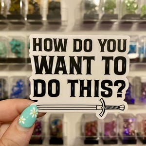 May include: A white sticker featuring the black text "HOW DO YOU WANT TO DO THIS?" and a sword illustration. The sticker is held up against a backdrop of small, colourful display items.