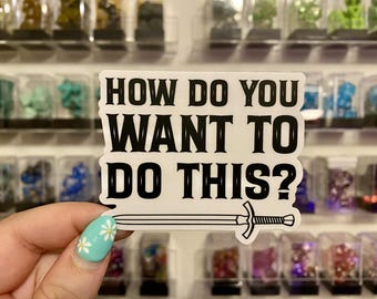 May include: A white sticker with the text "HOW DO YOU WANT TO DO THIS?" in black, with a sword illustration below. The sticker is held in front of a display of small, colorful objects.