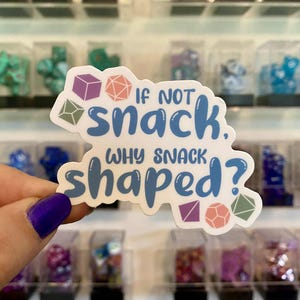 May include: A white sticker with the text "If not snack, why snack shaped?" in blue, with colorful geometric shapes. The sticker is held in front of a display of dice.