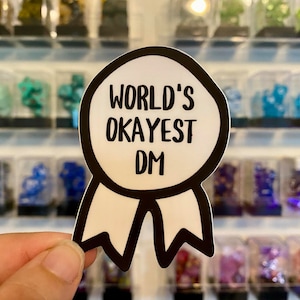 May include: A white sticker with a black outline in the shape of a ribbon. The ribbon has a circle in the center with the text "WORLD'S OKAYEST DM" in black.