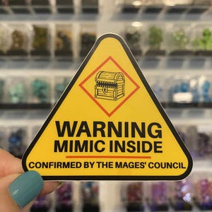 Puede incluir: Una pegatina triangular amarilla con el texto "WARNING MIMIC INSIDE" y una ilustración de un cofre. El texto "CONFIRMED BY THE MAGES' COUNCIL" está debajo. La pegatina se sostiene frente a un fondo borroso.