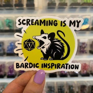 May include: A sticker featuring a black and white opossum with an open mouth, a yellow circle background, and the text "SCREAMING IS MY BARDIC INSPIRATION". The sticker also includes a black and white D20 die.