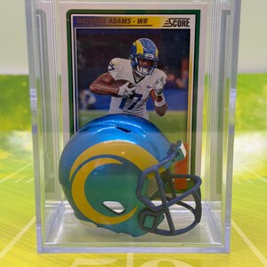 Puede incluir: Una vitrina de acrílico transparente que contiene una tarjeta coleccionable de fútbol y un casco en miniatura. La tarjeta presenta a Davante Adams - WR y el logotipo de Score. El casco es azul y amarillo, con el logotipo del equipo.