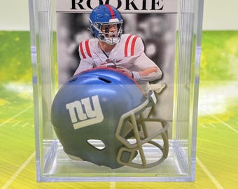 May include: A clear plastic display case with a miniature football helmet inside. The helmet is blue with a white "NY" logo and a brown face mask. The case also includes a card with a football player wearing a blue and red jersey. The card reads "Leaf Rookie".