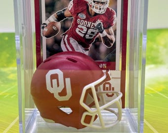May include: A miniature Oklahoma Sooners football helmet and a trading card are encased in a clear plastic display case. The helmet is crimson with a white facemask and the OU logo. The card features a player in a Sooners uniform.
