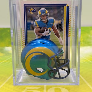 Puede incluir: Una tarjeta de fútbol y un casco en miniatura se exhiben en una caja de acrílico transparente. La tarjeta presenta a un jugador con uniforme azul y amarillo. El casco es azul y verde con un logotipo amarillo.