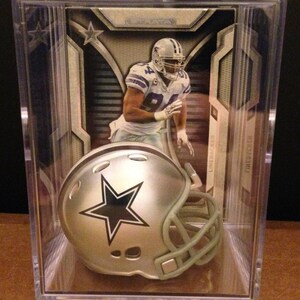 May include: A silver football helmet with a blue star on the side, displayed in a clear plastic case with a football player card in the background. The card features a player in a blue and white uniform with the number 81 on the front.