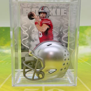 May include: A sports trading card featuring a football player in a red jersey throwing a ball, labeled PRIZED ROOKIE, displayed in a clear plastic case with a miniature silver football helmet with star decals positioned in front.