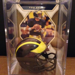 May include: A clear display case containing a Tom Brady football card and a miniature football helmet. The helmet is brown and yellow, with a black facemask. The card features Tom Brady in a Michigan Wolverines uniform.