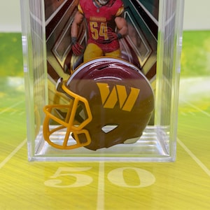 May include: A miniature burgundy and gold Washington Commanders football helmet with a yellow facemask, displayed in a clear plastic protective case in front of a sports trading card featuring a player in a burgundy jersey with the number 54.