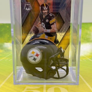 May include: A miniature Pittsburgh Steelers football helmet and a football card are displayed inside a clear plastic case. The helmet is olive green with the Steelers logo. The card features a player in a black and gold uniform.