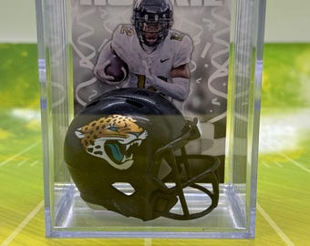 Può includere: Un casco da football americano in miniatura di colore nero e turchese con il logo dei Jacksonville Jaguars è esposto in una custodia di plastica trasparente. La custodia include anche una carta con un giocatore di football americano che indossa una maglia bianca con il numero 25 e la scritta "PRIZED ROOKIE" su di essa.