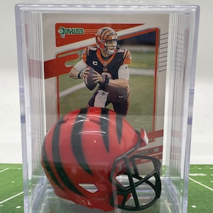 May include: A clear plastic display case containing a 2021 Donruss football trading card of Joe Burrow and a miniature orange football helmet with black tiger stripes and a black face mask. The card features the player in a black uniform.