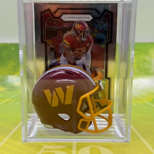 May include: A miniature burgundy and gold football helmet with a yellow face mask, displayed in a clear plastic protective case in front of a sports trading card featuring a Washington Commanders player with the text COMMANDERS at the top.