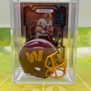 May include: A miniature burgundy and gold football helmet with a yellow face mask, displayed in a clear acrylic case in front of a trading card featuring a football player in a white jersey with the text Redskins printed at the top.