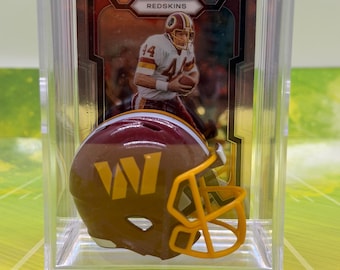Könnte beinhalten: Ein Miniatur-Footballhelm in Burgunderrot und Gold mit gelbem Gesichtsschutz, präsentiert in einer durchsichtigen Acrylhülle vor einer Sammelkarte, die einen Spieler im weißen Trikot mit dem Text Redskins am oberen Rand zeigt.