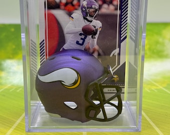 May include: A purple and gold miniature football helmet with a Minnesota Vikings logo is displayed in a clear plastic case with a football card featuring a player in a Vikings uniform.
