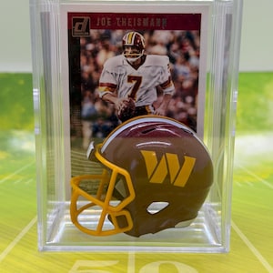 Op de afbeelding: Een Donruss Joe Theismann football-verzamelkaart tentoongesteld achter een miniatuur bordeauxrode en gouden footballhelm met een geel gezichtsmasker, alles verpakt in een rechthoekige beschermende behuizing van doorzichtig plastic.