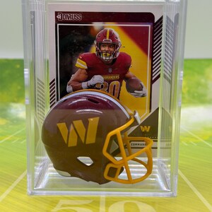May include: A Donruss football trading card featuring a Washington Commanders player in a burgundy and gold uniform, displayed in a clear plastic case with a miniature burgundy and gold football helmet with a yellow facemask in front.