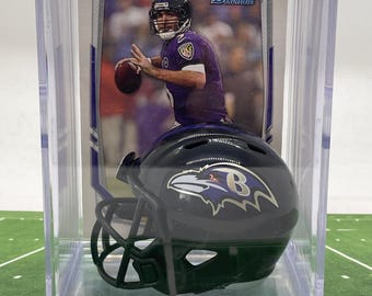 May include: A clear plastic display case with a miniature Baltimore Ravens football helmet and a Bowman trading card featuring a player in a purple jersey. The card has the text "Bowman" on it.