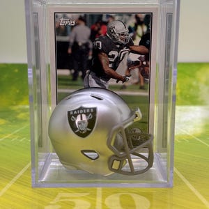 May include: A silver miniature football helmet with a black Raiders logo and a clear plastic display case. The case also includes a Topps trading card of a football player wearing a Raiders uniform.