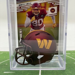 May include: A clear plastic display case containing a miniature football helmet. The helmet is burgundy with a gold "W" on the side. The case also includes a trading card featuring a football player in a burgundy uniform with the number "90" on the front. The card reads "MONTEZ SWEAT".
