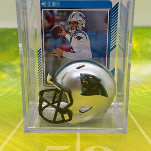 May include: A silver football helmet with a black facemask and a blue and black Carolina Panthers logo. The helmet is displayed in a clear plastic case with a trading card featuring a football player in the background.