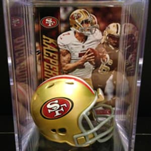 May include: A miniature gold San Francisco 49ers football helmet with a red stripe and logo, displayed inside a clear rectangular plastic protective case. Behind the helmet is a trading card featuring a football player in a white jersey.