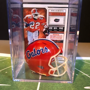 Könnte beinhalten: Eine transparente Acryl-Vitrine mit einem Football-Helm und einer Sammelkarte. Der Helm ist orange mit einem blau-weißen "Gators"-Logo. Die Karte zeigt einen Footballspieler in einem orangefarbenen Trikot mit der Nummer 22.
