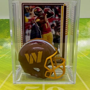 May include: A miniature brown and yellow football helmet with a W logo, displayed in a clear plastic case in front of a trading card featuring a football player in a burgundy and gold uniform catching a football with a W logo visible.