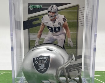 Może przedstawiać: Srebrny hełm do futbolu amerykańskiego z logo Raiders, wystawiony w przezroczystej plastikowej gablocie. Gablotka zawiera również kartę piłkarską gracza w stroju Raiders.