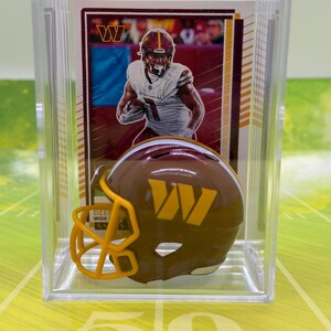 May include: A clear plastic display case containing a Washington Commanders football trading card and a miniature burgundy and gold football helmet. The card features a player in a white uniform, and the helmet displays a gold W logo on the side.