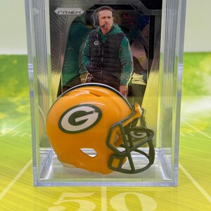 May include: A clear plastic case containing a football trading card and a miniature football helmet. The card features a man in a green jacket and black vest. The helmet is yellow and green with the letter "G".