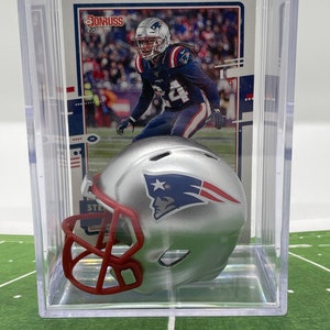May include: A silver and red miniature football helmet with a white star and the New England Patriots logo. The helmet is in a clear plastic display case with a football card featuring a player in a Patriots uniform.